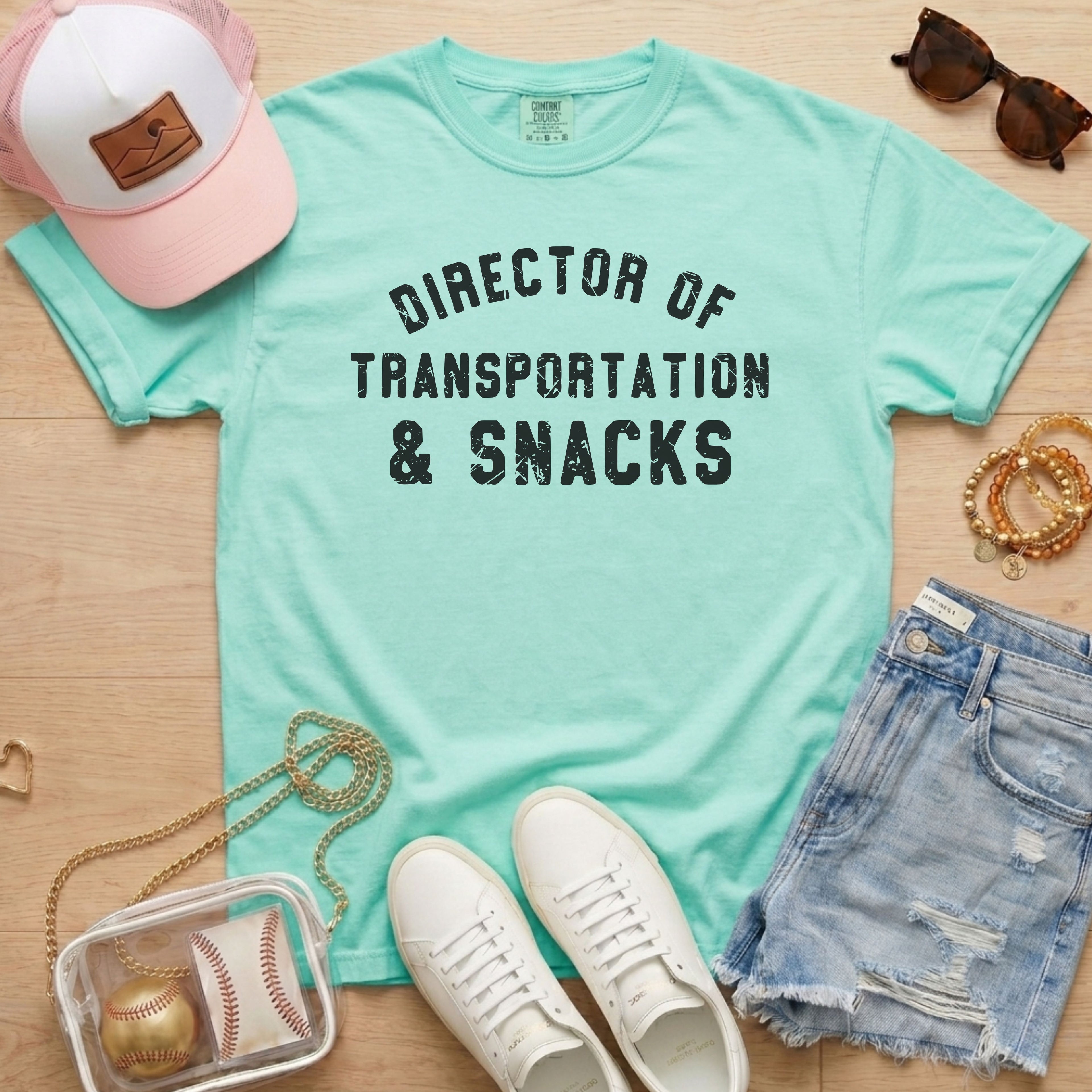Flat lay of a distressed mint green t-shirt with the slogan 'DIRECTOR OF TRANSPORTATION & SNACKS' and tortoise sunglasses.