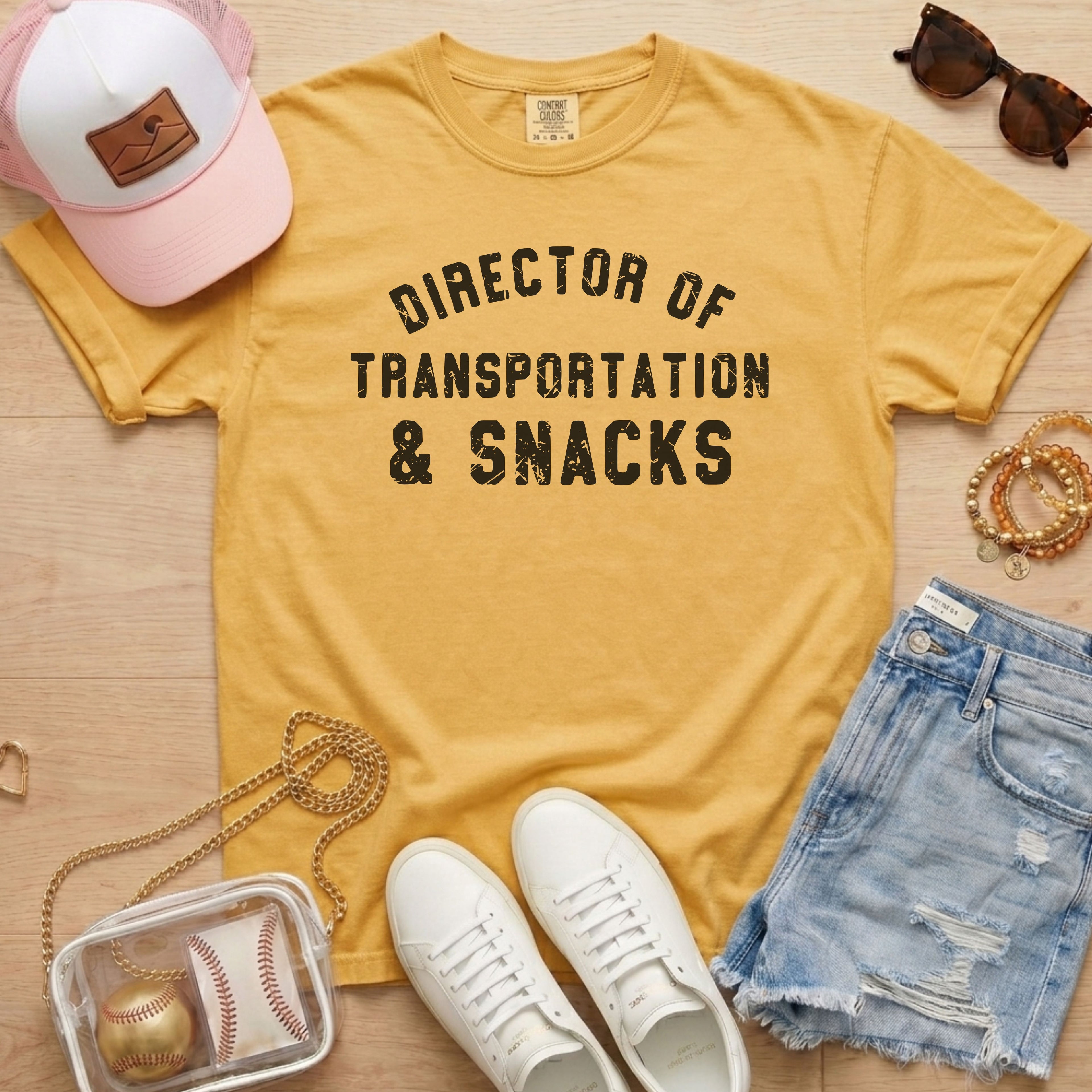 Flat lay of a distressed mustard yellow t-shirt with the slogan 'DIRECTOR OF TRANSPORTATION & SNACKS' and clear stadium bag.
