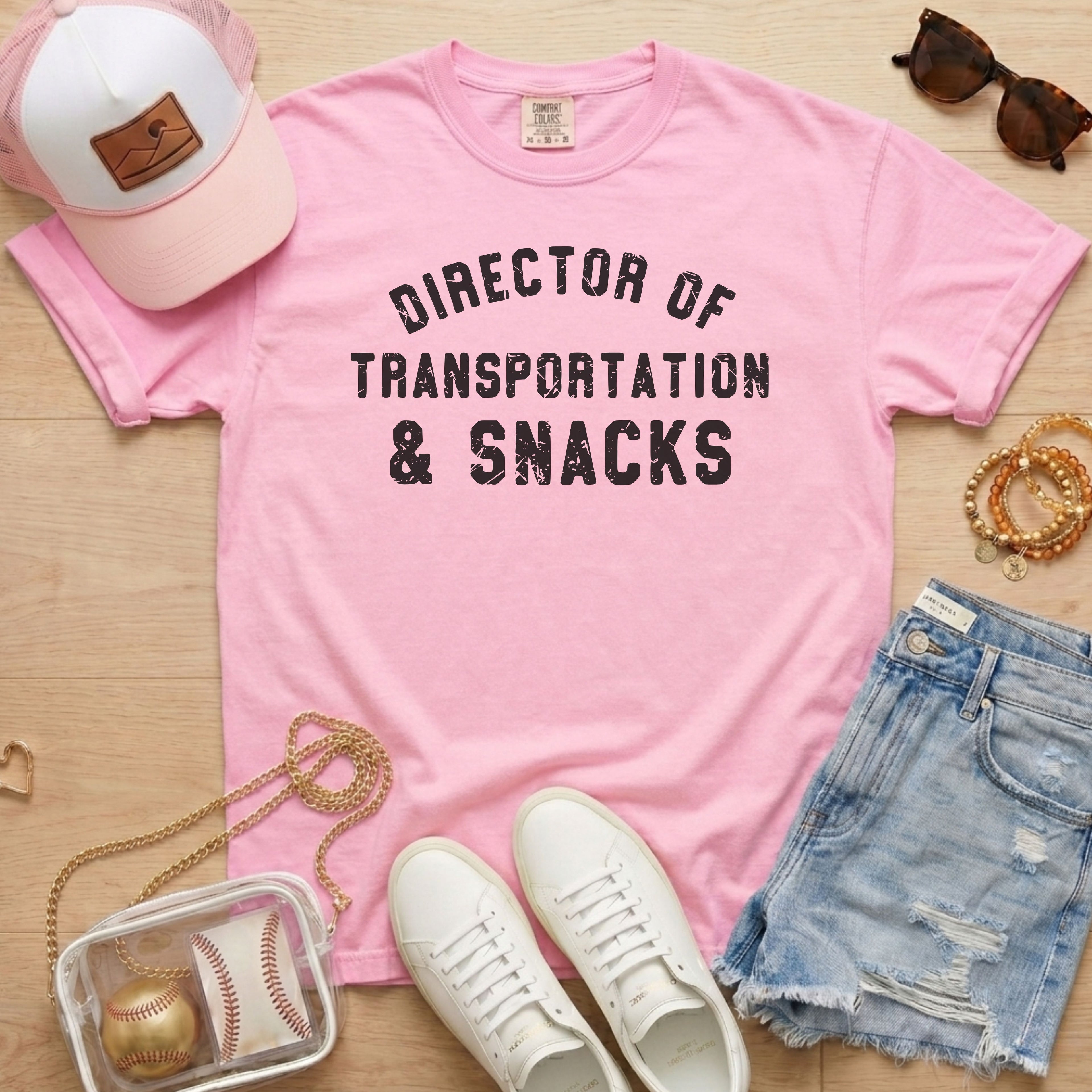 Flat lay of a distressed soft pink t-shirt with the slogan 'DIRECTOR OF TRANSPORTATION & SNACKS' and white sneakers.