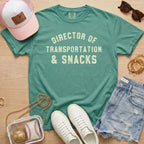 Flat lay of a distressed teal green t-shirt with the slogan 'DIRECTOR OF TRANSPORTATION & SNACKS' and gold bracelets.