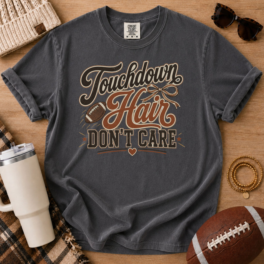 A dark gray graphic t-shirt laid flat on a wooden surface, surrounded by lifestyle props including a football, sunglasses, a tumbler, and a beanie. The t-shirt features a distressed graphic that reads 'Touchdown Hair DON'T CARE' with a football and bowtie illustration.
