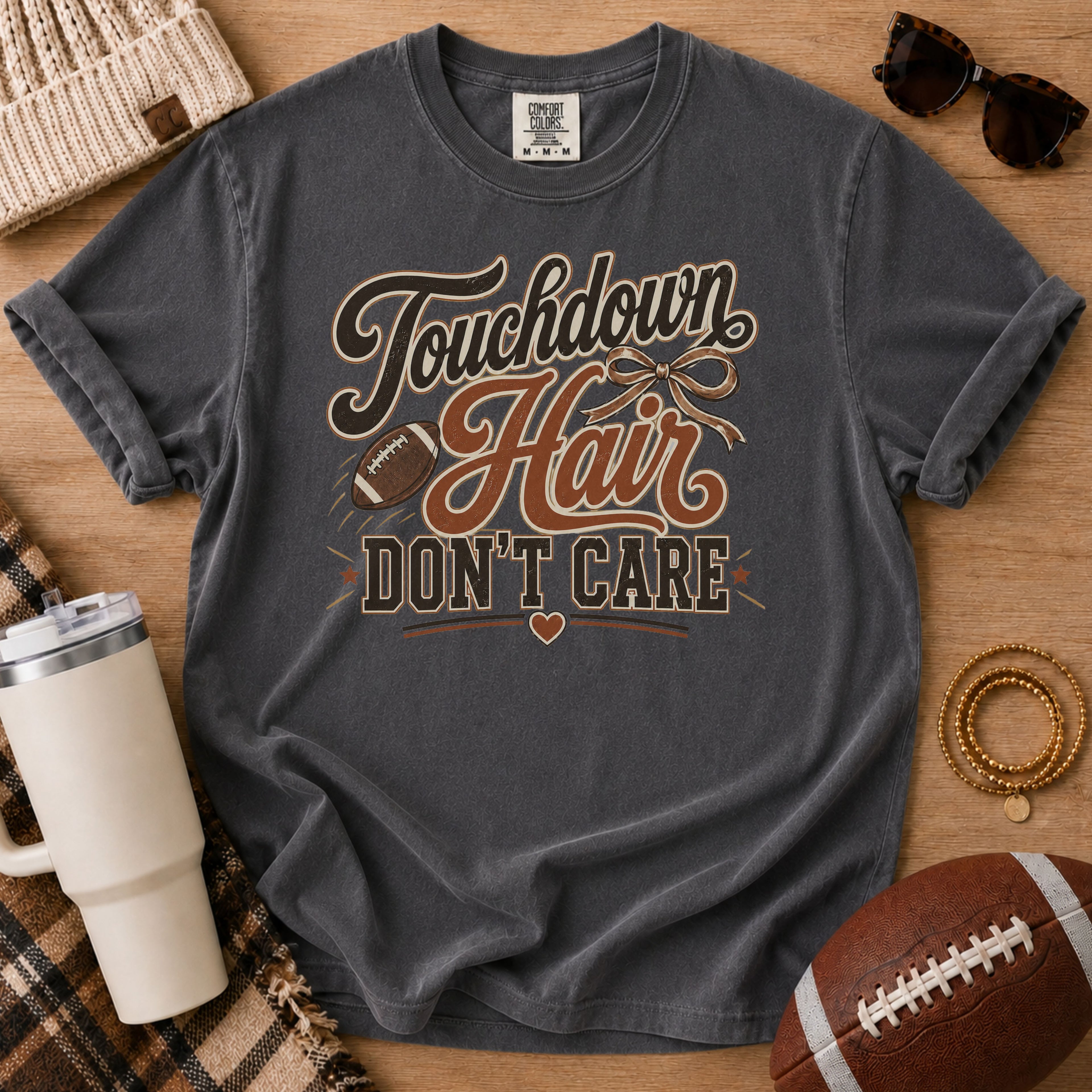 A dark gray graphic t-shirt laid flat on a wooden surface, surrounded by lifestyle props including a football, sunglasses, a tumbler, and a beanie. The t-shirt features a distressed graphic that reads 'Touchdown Hair DON'T CARE' with a football and bowtie illustration.