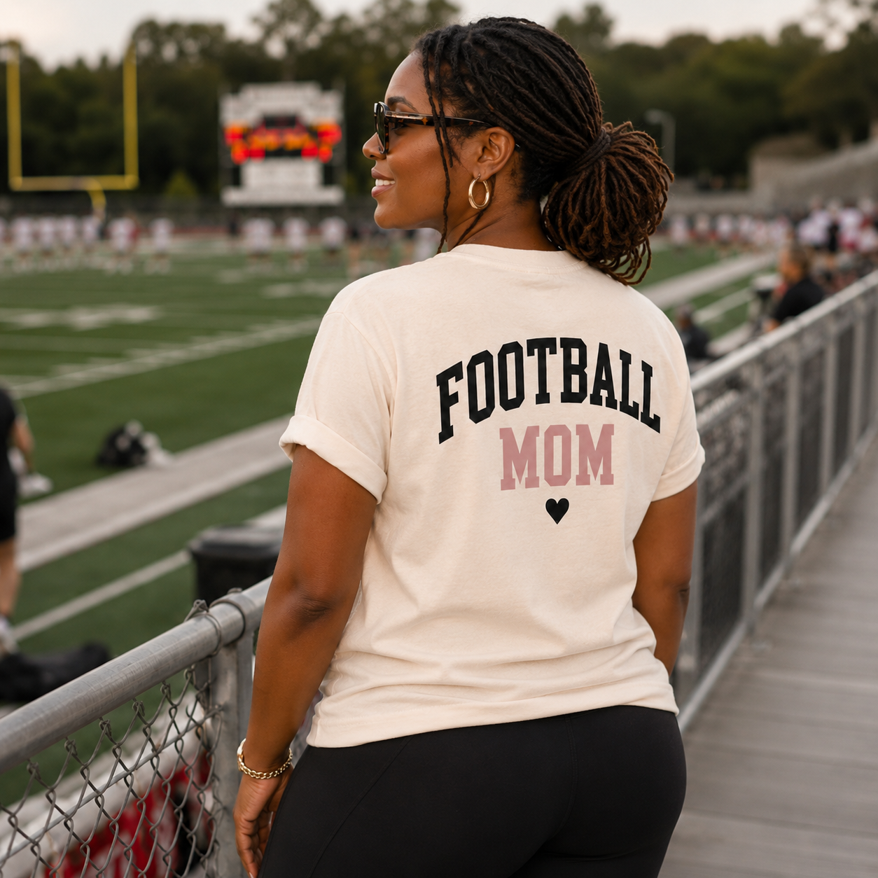 Woman wearing the FOOTBALL MOM graphic shirt in cream, Comfort Colors Ivory, at a football field with the back design visible.