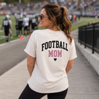 A woman standing on a football field walkway wearing a cream-colored t-shirt with 'FOOTBALL MOM' printed on the back in black and pink lettering.