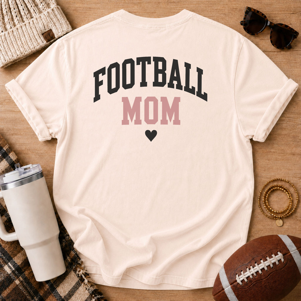 Woman wearing the FOOTBALL MOM graphic shirt in cream, Comfort Colors Ivory, at a football field with the back design visible.