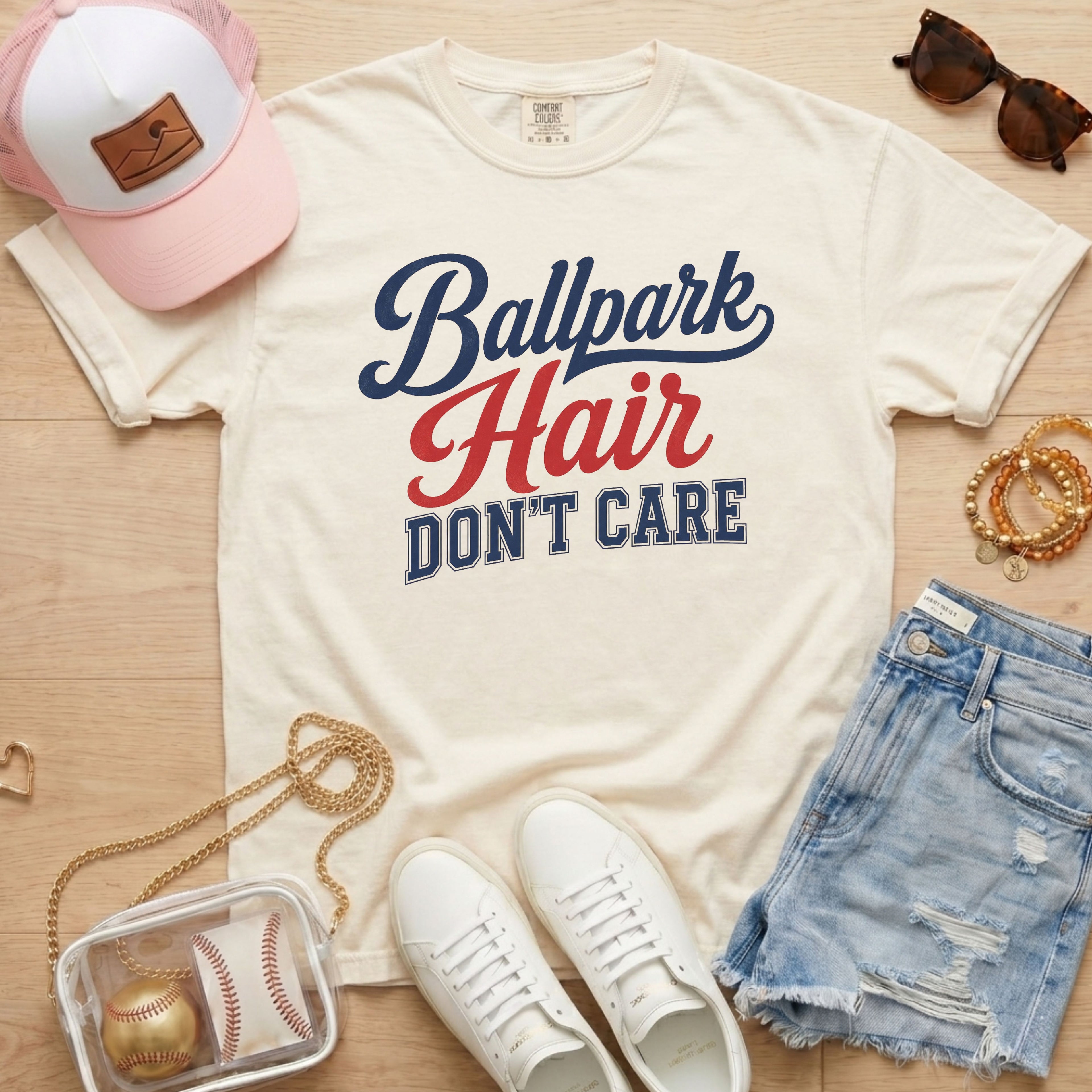 Ivory Ballpark Hair Don’t Care funny baseball mom shirt styled with denim shorts, sneakers, pink hat, clear baseball bag, and sideline accessories.
