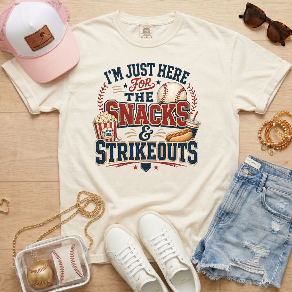 Ivory Snacks & Strikeouts funny baseball game day shirt styled with denim shorts, white sneakers, pink hat, sunglasses, jewelry, and clear baseball bag.