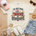 Ivory Snacks & Strikeouts funny baseball game day shirt styled with denim shorts, white sneakers, pink hat, sunglasses, jewelry, and clear baseball bag.