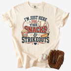 Ivory Comfort Colors Snacks & Strikeouts funny baseball mom
shirt displayed with a baseball and glove, featuring a vintage snack stand and strikeouts design.