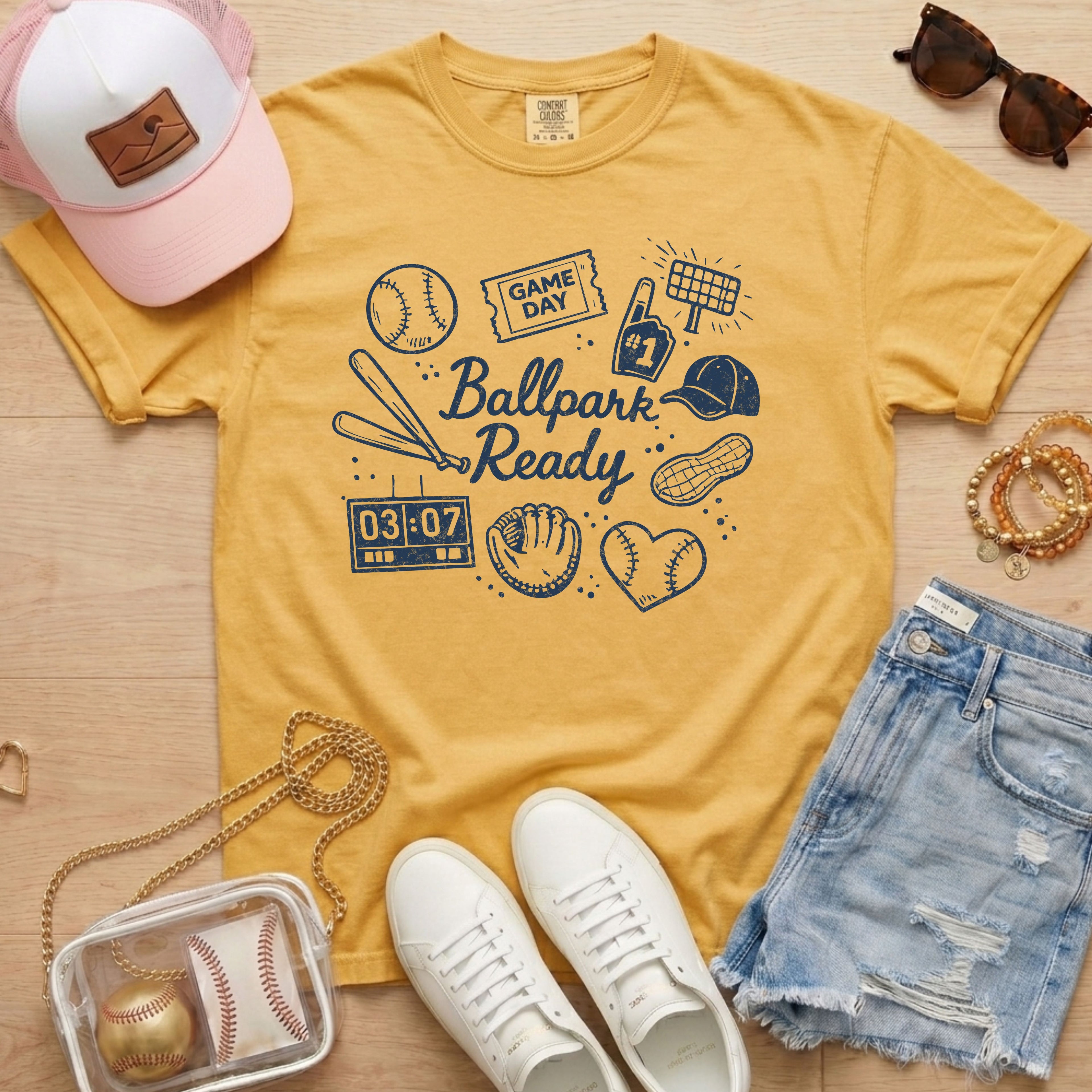 Yellow t-shirt with 'Ballpark Ready' design, white sneakers, denim shorts, pink cap, and baseball glove on a wooden surface.