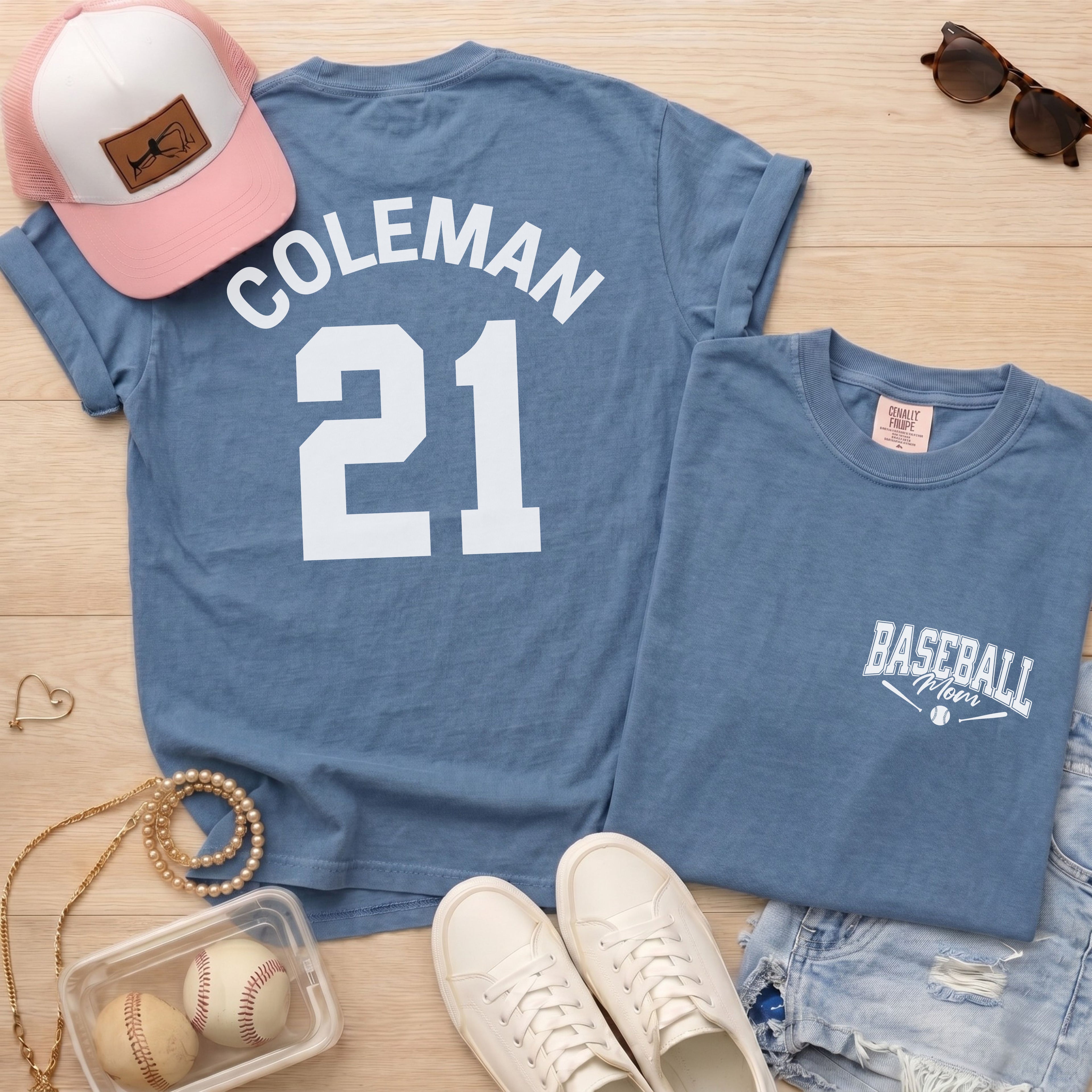 Blue t-shirt with 'Coleman 21' design, pink cap, sunglasses, and baseballs on a wooden surface.