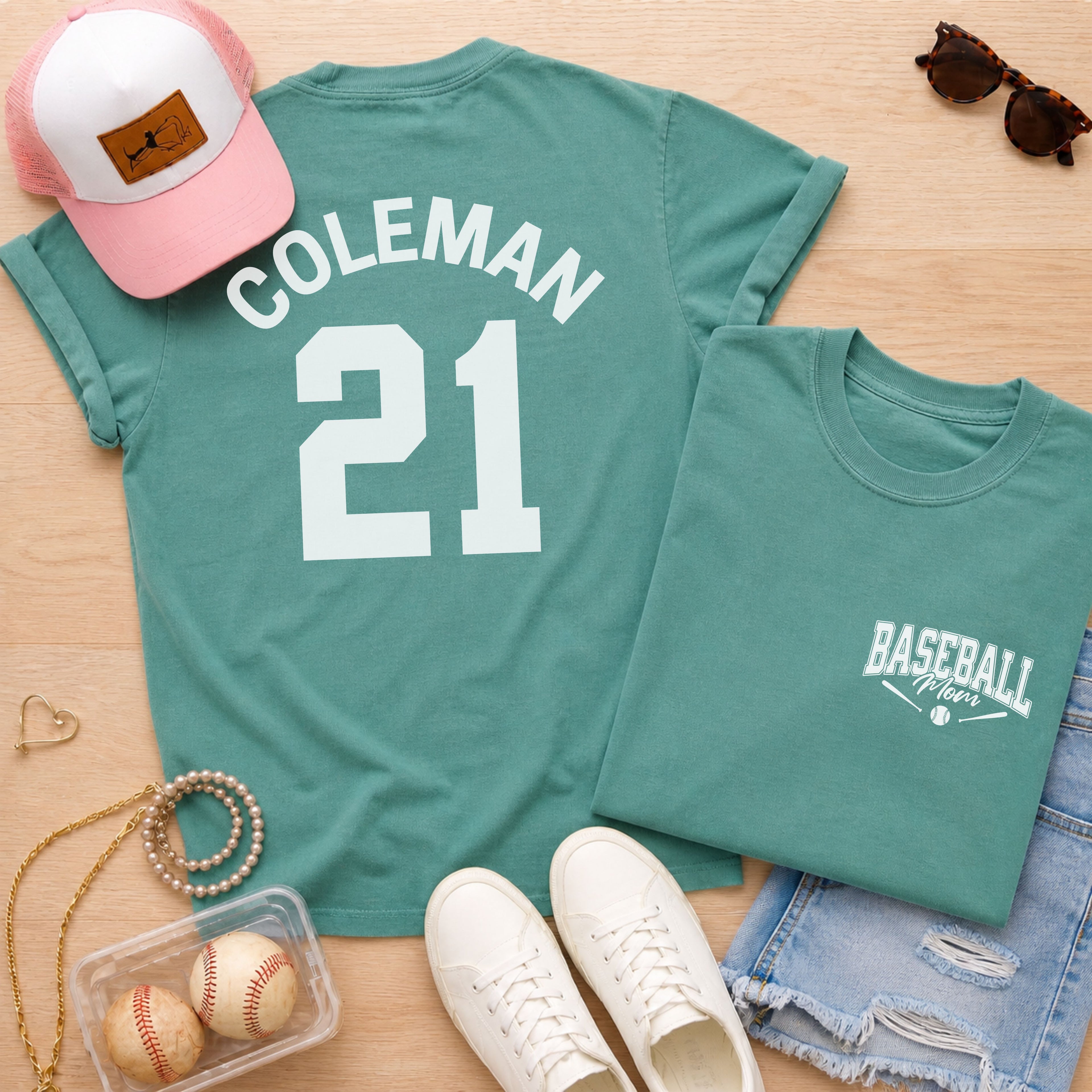 Teal t-shirt with 'Coleman 21' design, pink cap, sunglasses, and baseballs on a wooden surface.