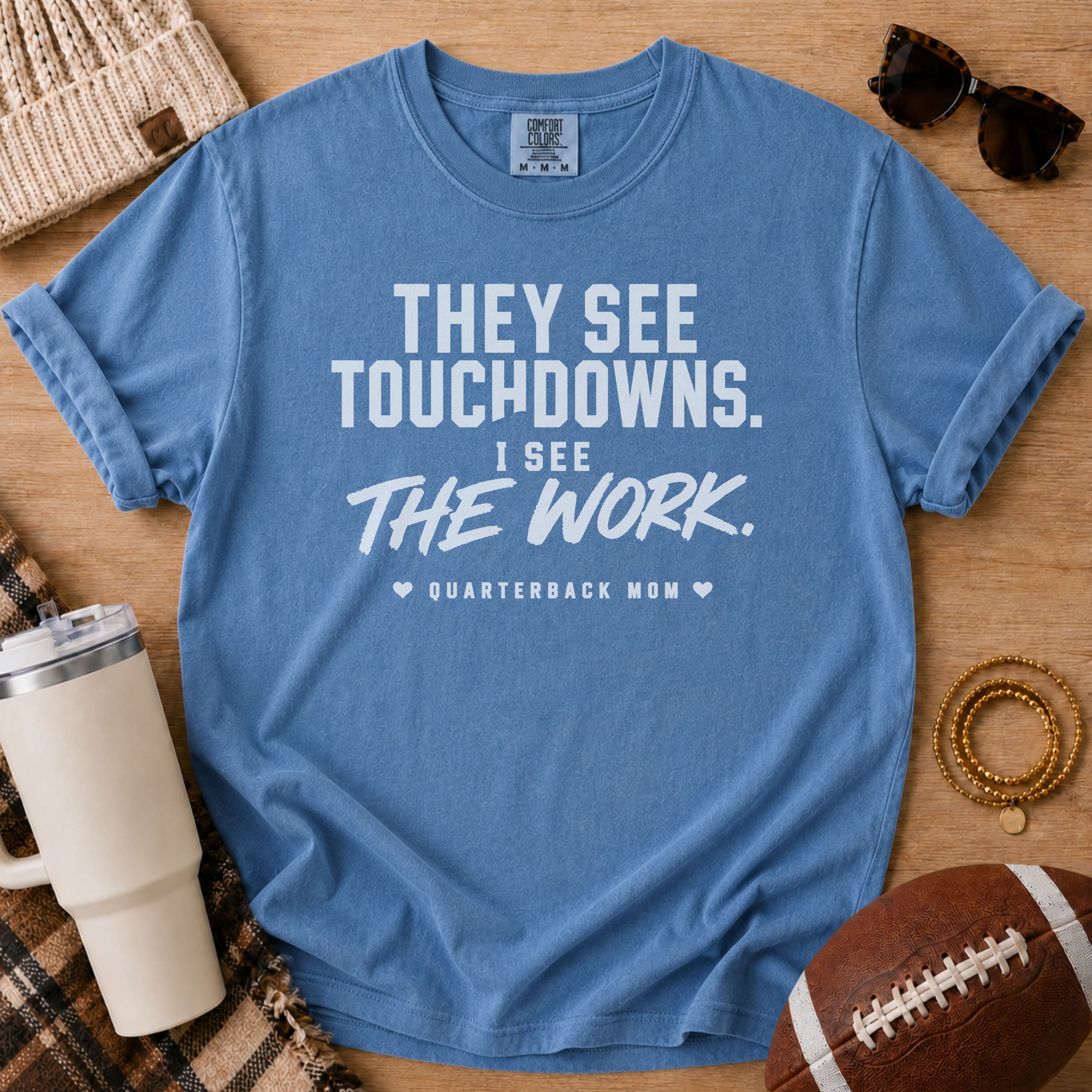 A blue short-sleeve graphic T-shirt laid flat on a wooden surface with lifestyle props including sunglasses, a beanie, a tumbler, a football, and bracelets. The shirt has white text reading, 'THEY SEE TOUCHDOWNS.