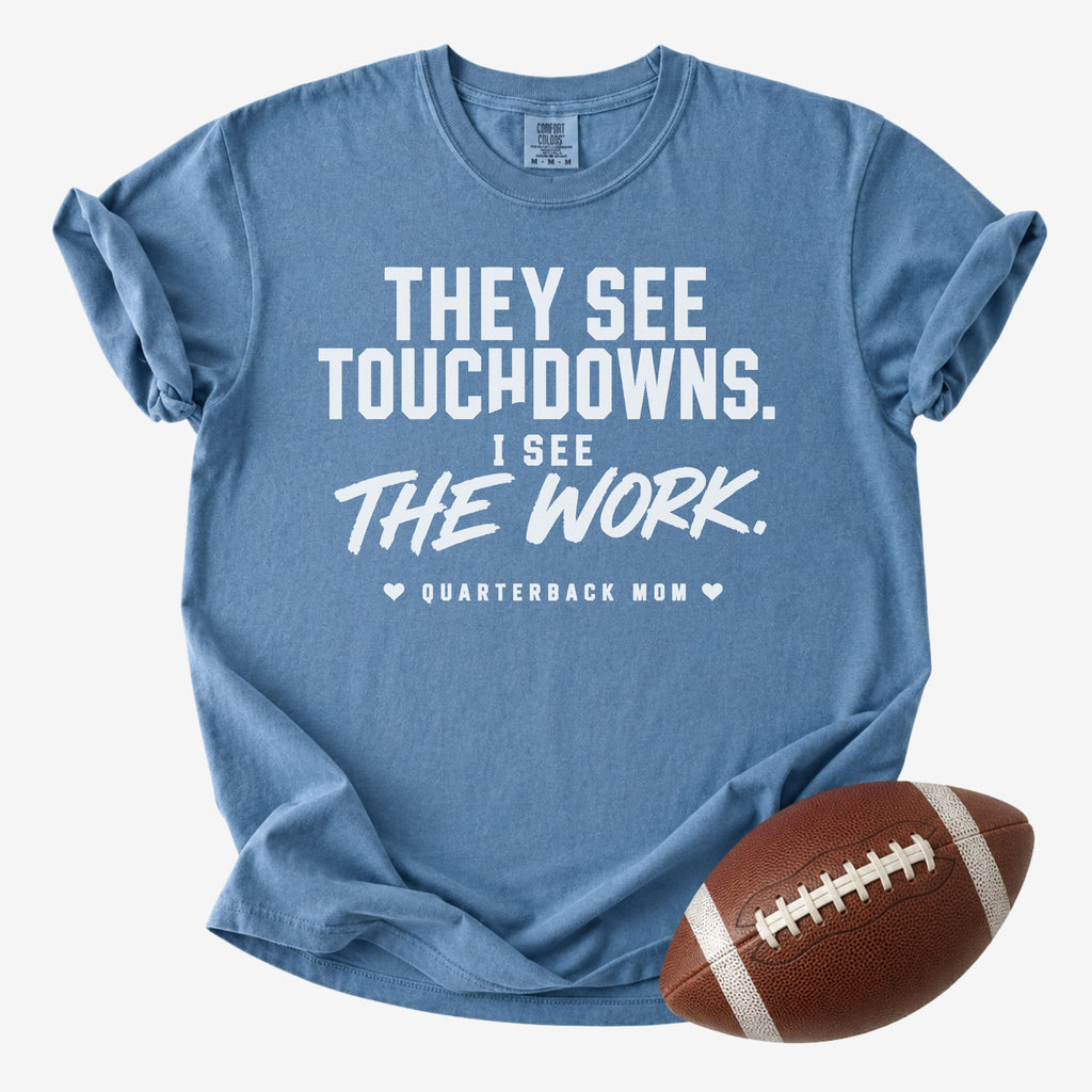 A heathered blue short-sleeve t-shirt with a white printed graphic that reads 'THEY SEE TOUCHDOWNS. I SEE THE WORK.