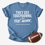 A heathered blue short-sleeve t-shirt with a white printed graphic that reads 'THEY SEE TOUCHDOWNS. I SEE THE WORK.