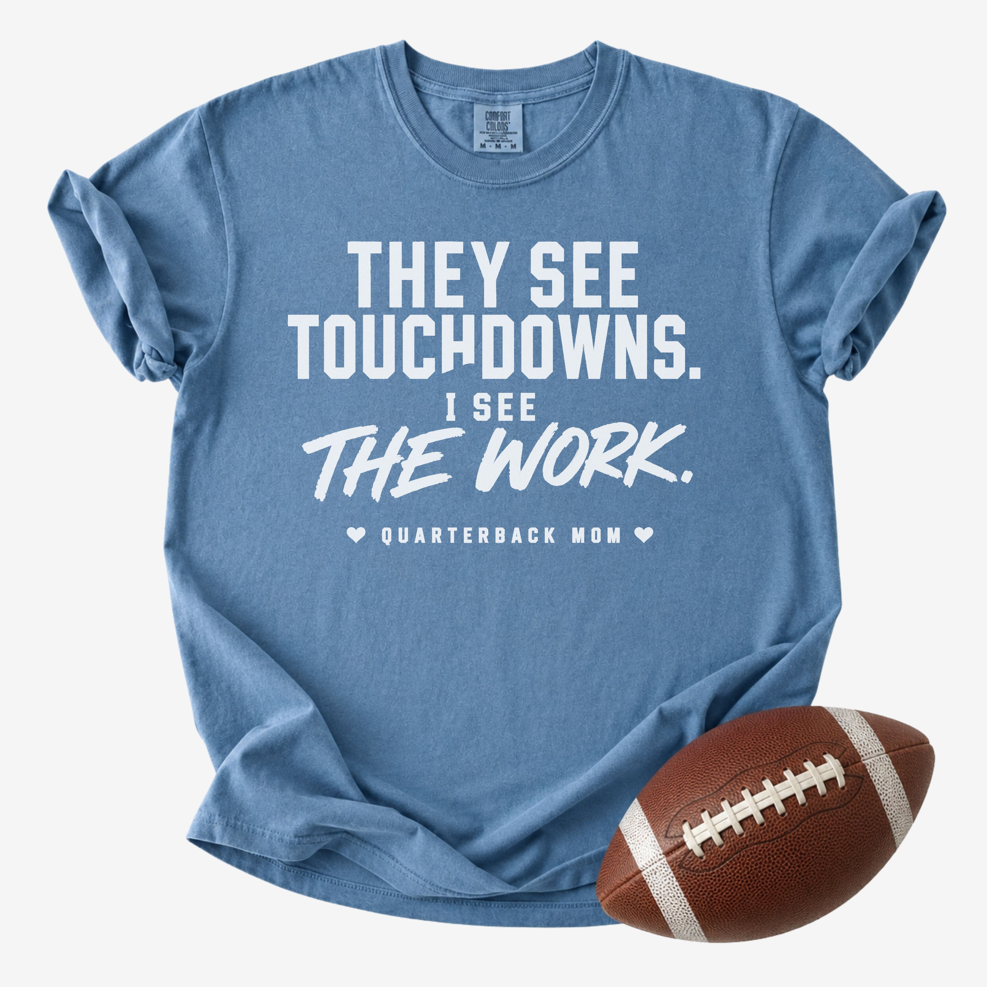 A heathered blue short-sleeve t-shirt with a white printed graphic that reads 'THEY SEE TOUCHDOWNS. I SEE THE WORK.