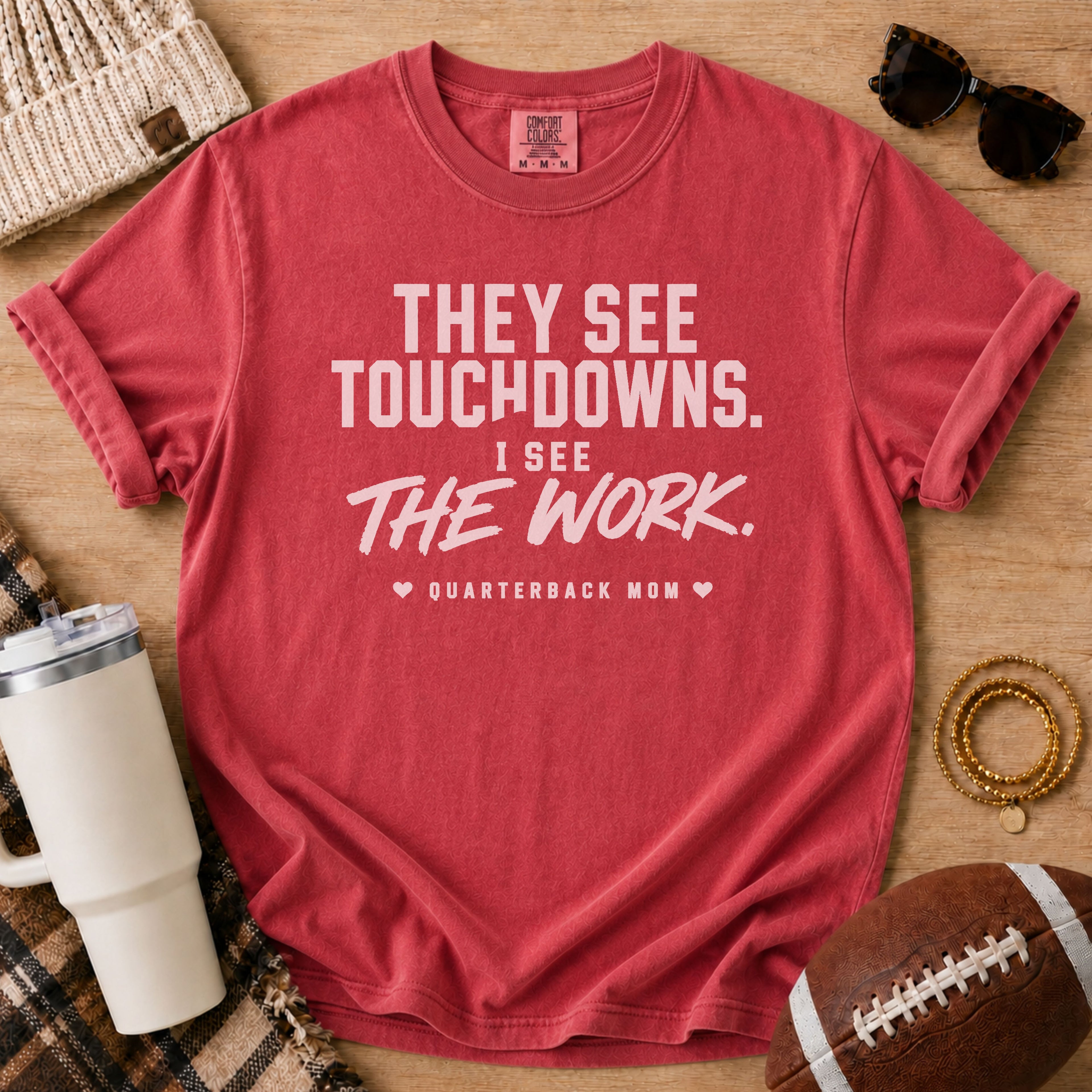 A red short-sleeve graphic T-shirt laid flat on a wooden surface with football-themed props, including a football, sunglasses, a tumbler, a knit hat, and bracelets. The shirt has white text reading, 'THEY SEE TOUCHDOWNS.