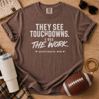 A brown short-sleeve graphic T-shirt laid flat on a wooden surface with football-themed props, including a football, sunglasses, a beanie, a tumbler, and bracelets. The shirt has large white text reading “THEY SEE TOUCHDOWNS.