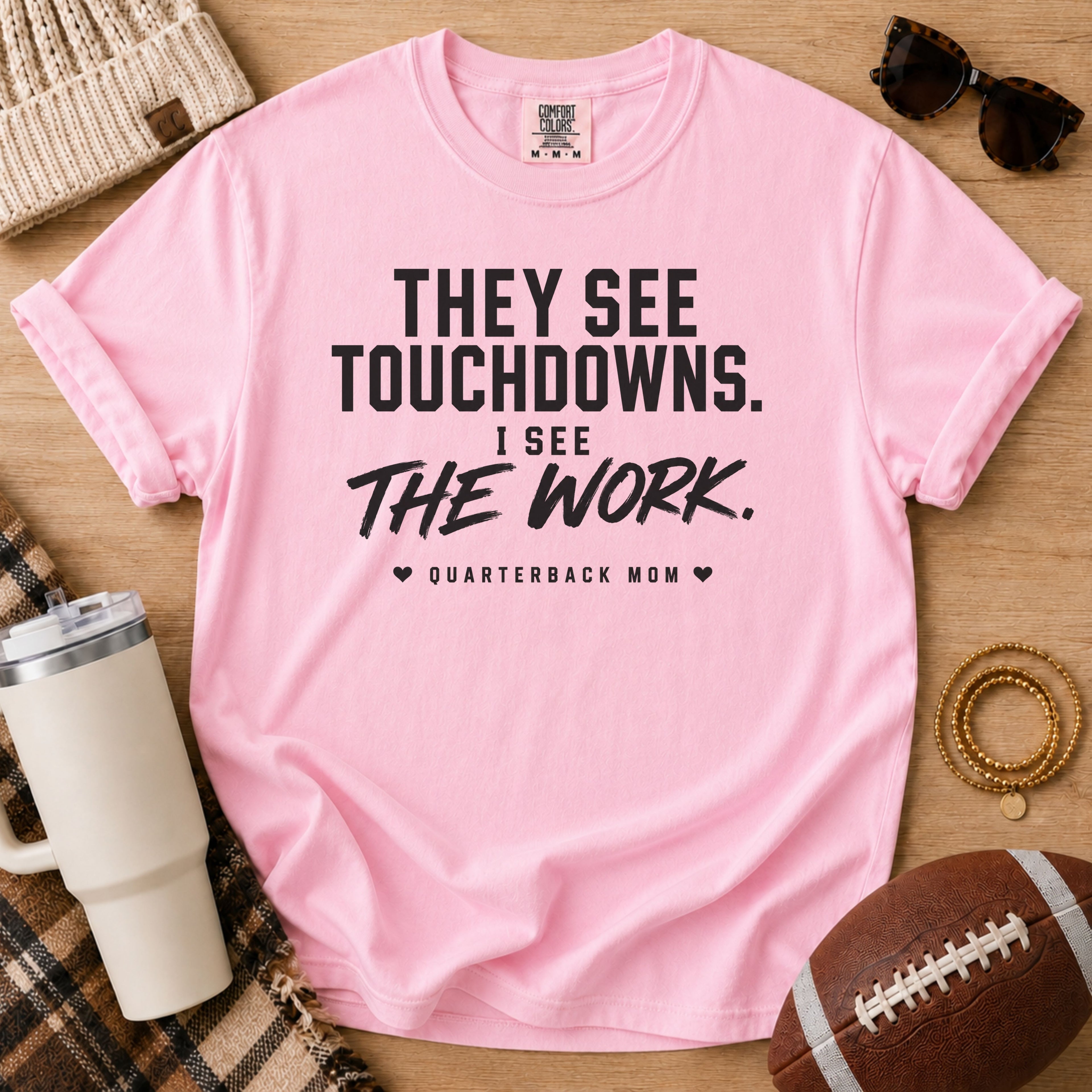 A pink short-sleeve graphic T-shirt laid flat on a wooden surface with sports-themed props including sunglasses, a beanie, a tumbler, a football, and bracelets. The shirt has bold black text reading 'THEY SEE TOUCHDOWNS.
