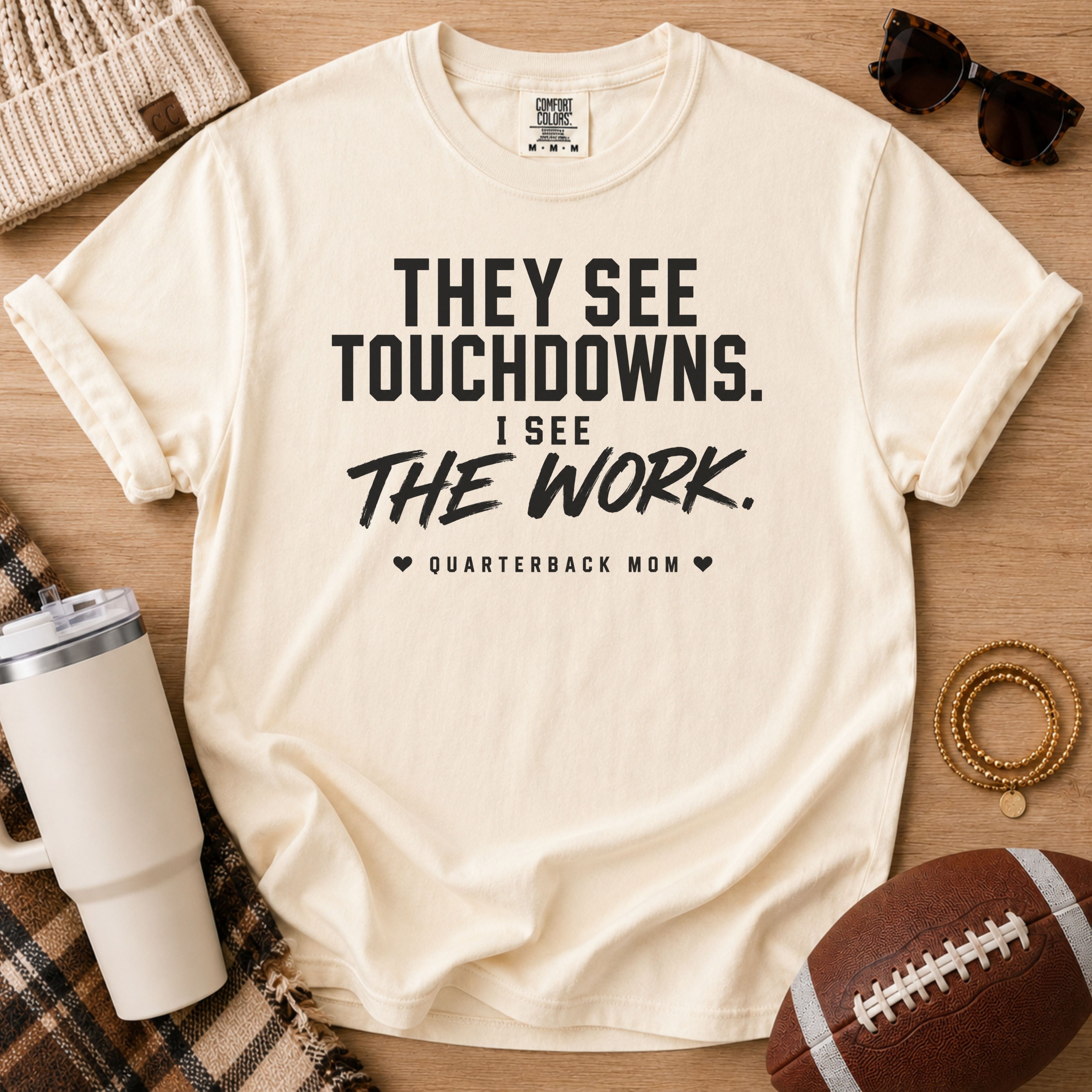 A cream-colored short-sleeve t-shirt with rolled sleeves, featuring the text 'THEY SEE TOUCHDOWNS. I SEE THE WORK.