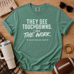 A sage green graphic t-shirt with white text reading 'THEY SEE TOUCHDOWNS. I SEE THE WORK.