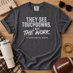 A dark charcoal gray graphic t-shirt laid flat on a wooden surface, surrounded by lifestyle props including a beige knit beanie, sunglasses, a cream tumbler, a football, and a plaid blanket. The t-shirt features the text 'THEY SEE TOUCHDOWNS.