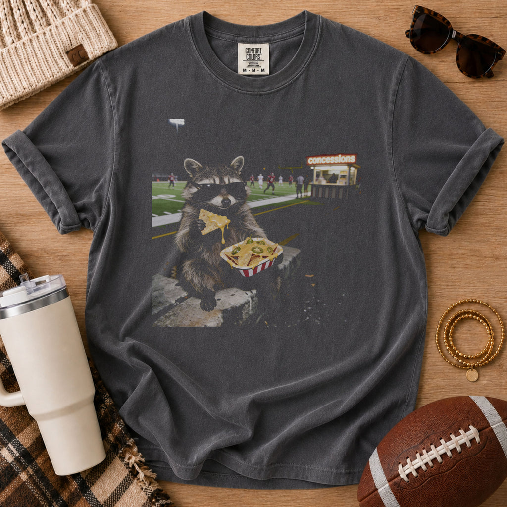 Charcoal gray football game day graphic tee, Comfort Colors Pepper, styled in a flat lay with a raccoon eating nachos near a football field and a football beside the shirt.