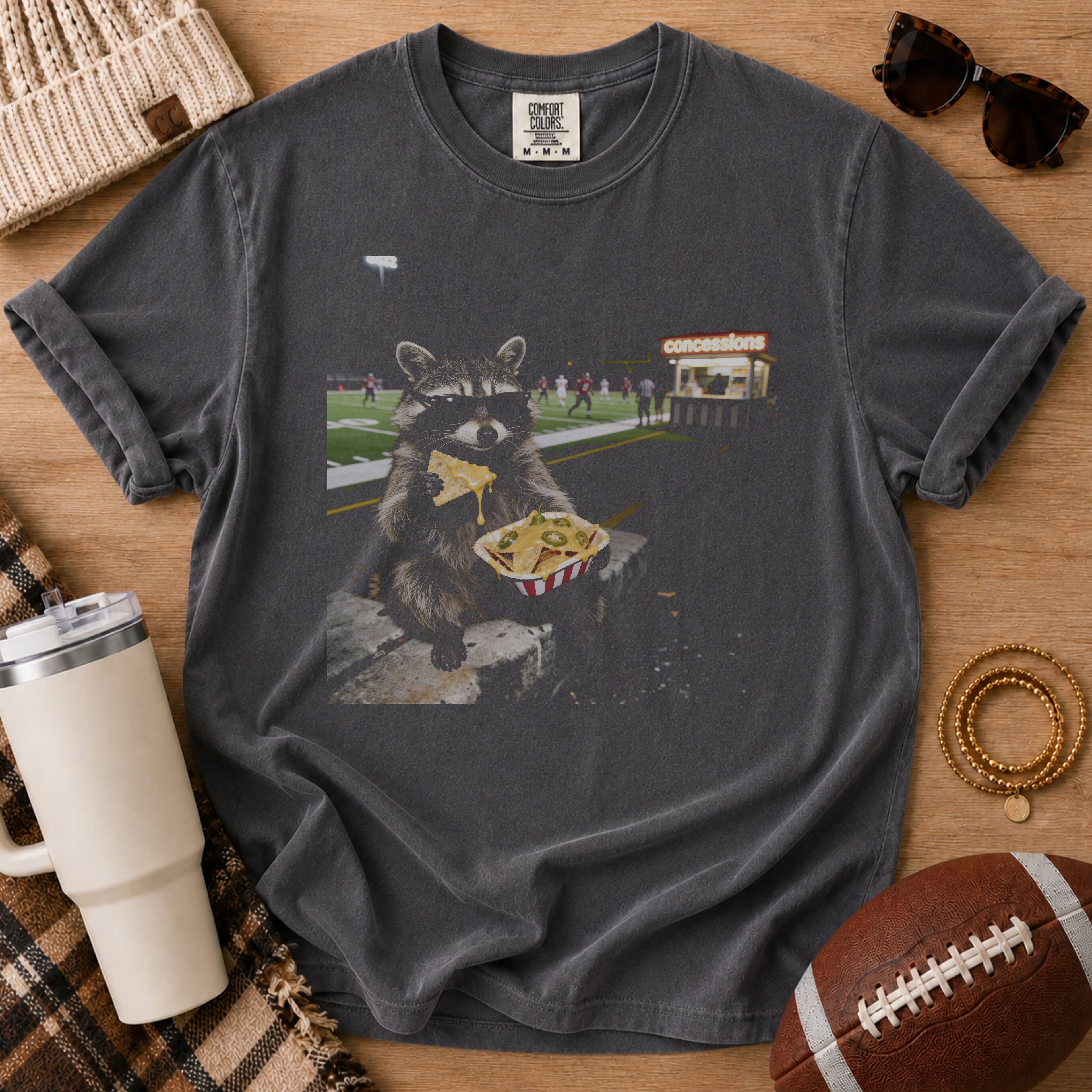 Charcoal gray football game day graphic tee, Comfort Colors Pepper, styled in a flat lay with a raccoon eating nachos near a football field and a football beside the shirt.
