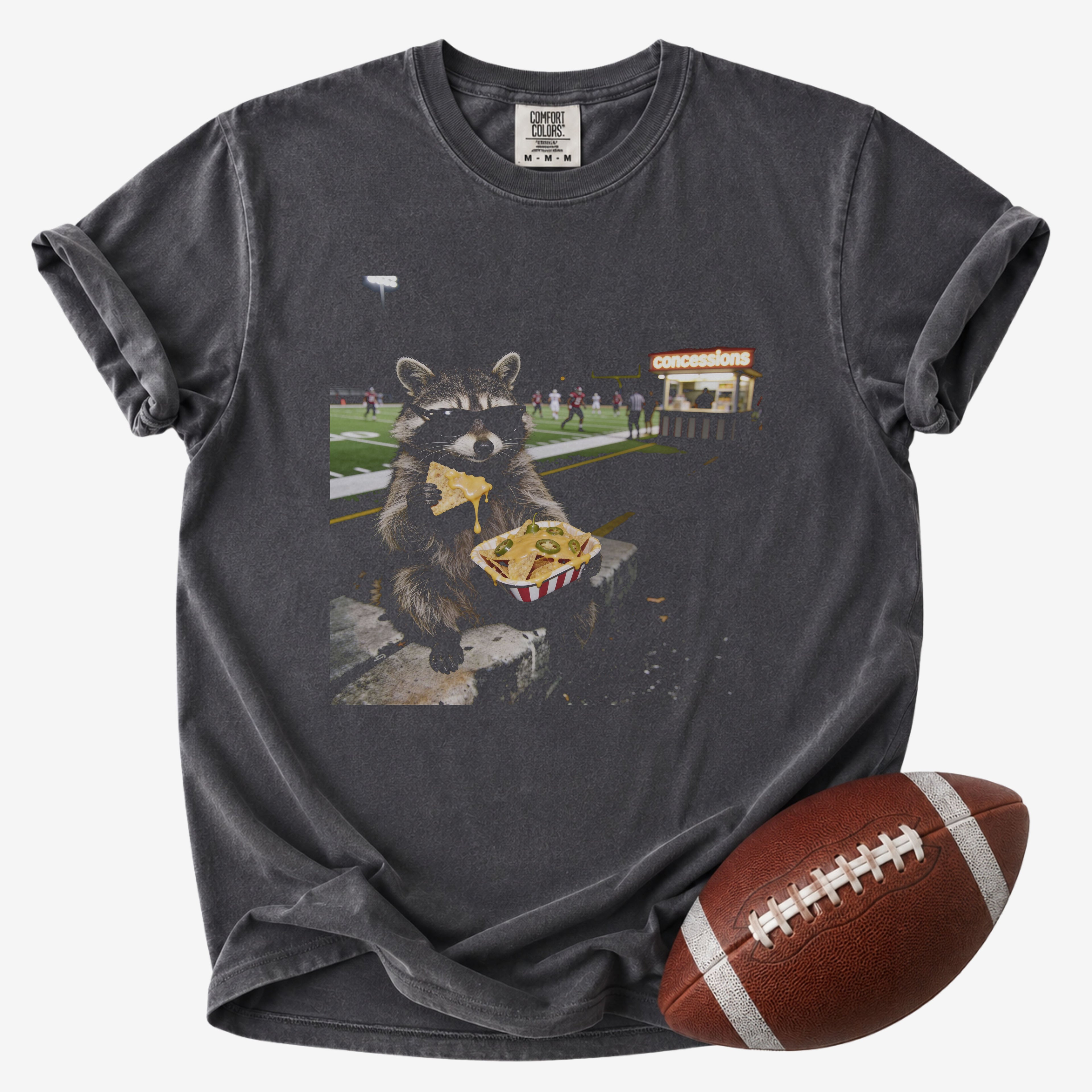 Funny football mom graphic T-shirt in charcoal gray, Comfort Colors Pepper, featuring a sunglasses-wearing raccoon eating nachos beside a football field and concession stand.