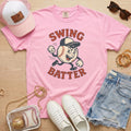 Pink t-shirt with 'Swing Batter' graphic, white sneakers, denim shorts, pink cap, and small bag on a wooden surface.