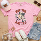 Pink t-shirt with 'Swing Batter' graphic, white sneakers, denim shorts, pink cap, and small bag on a wooden surface.