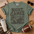 Vintage football graphic T-shirt in olive green, Comfort Colors Moss, styled in a game day flat lay with a distressed football and stadium-light design.