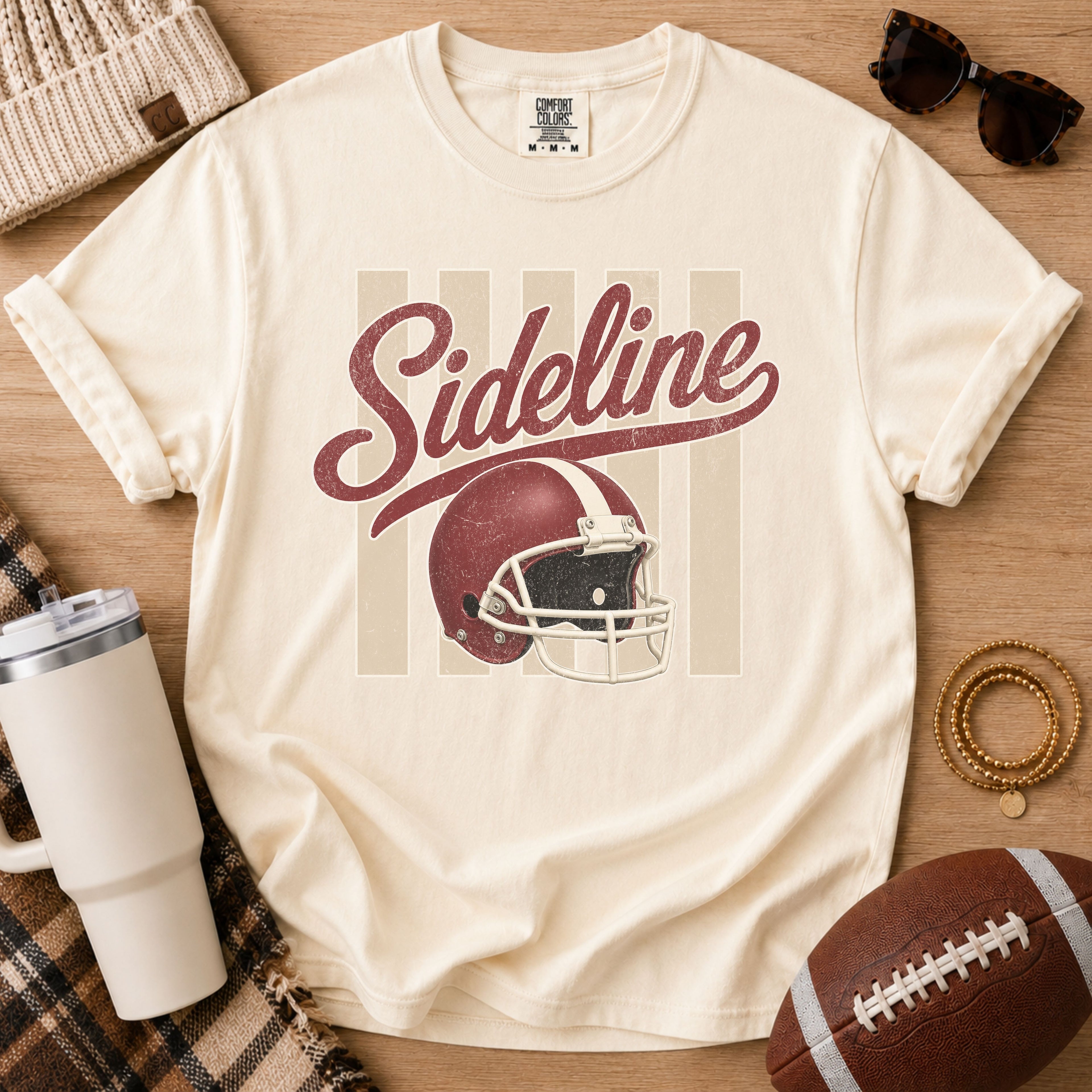 Cream football game day graphic tee, Comfort Colors Ivory, styled in a flat lay with a football and distressed “Sideline” helmet design.